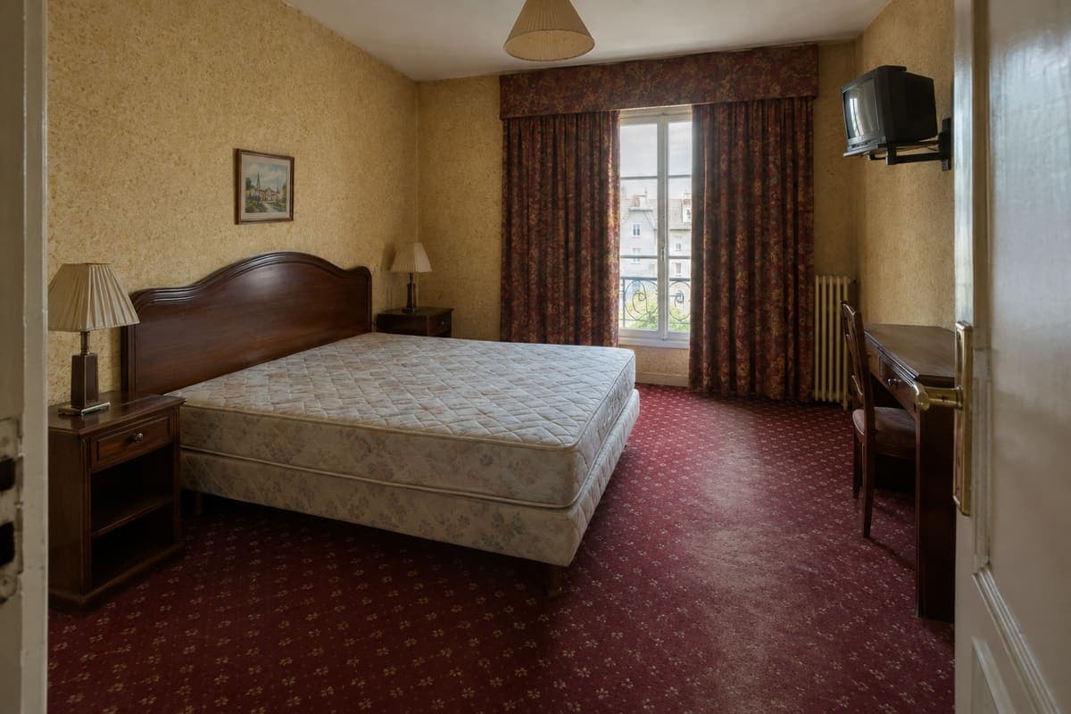 Chambre d’hôtel avant rénovation : moquette à motifs, mobilier et éclairage datés.